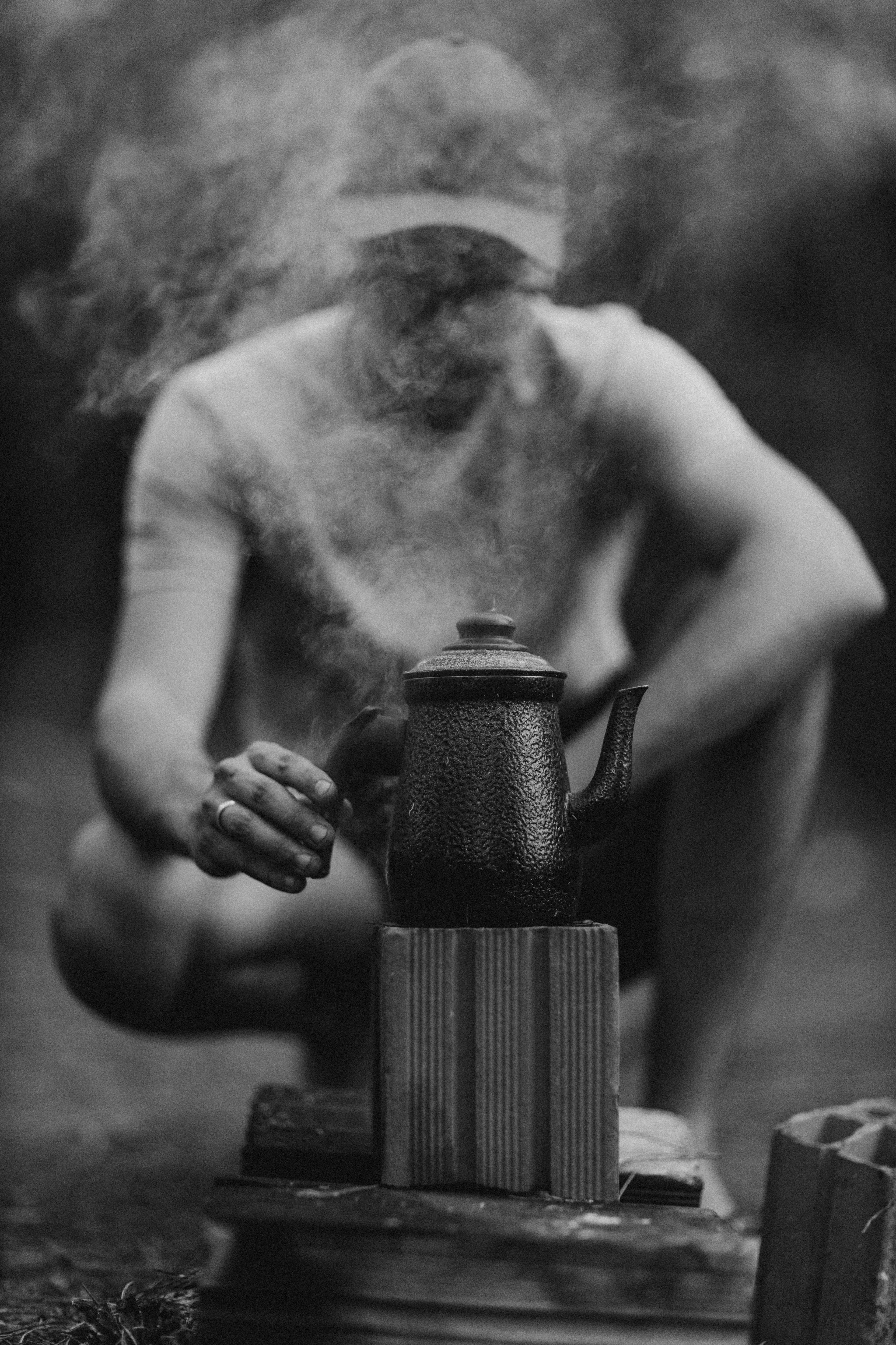 Atmospheric black and white photo of a man heating up a kettle outdoors.