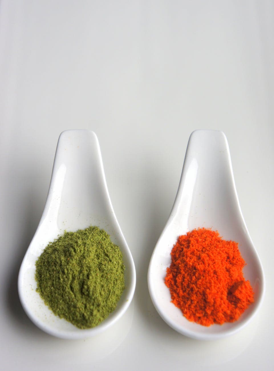 Two ceramic spoons with vibrant green and red spice powders on a neutral background.