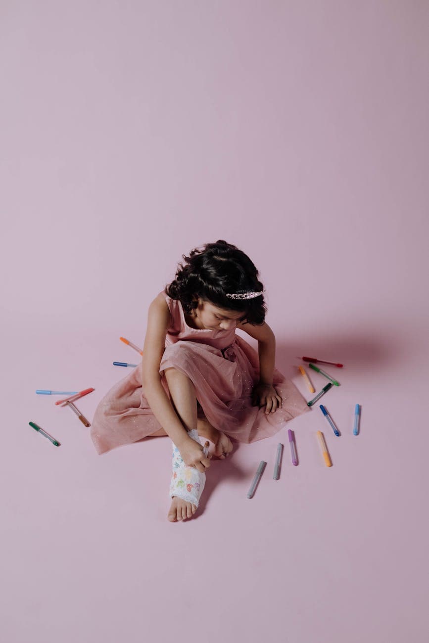 Girl in a pink dress with an orthopedic cast surrounded by colored markers on a pink background.