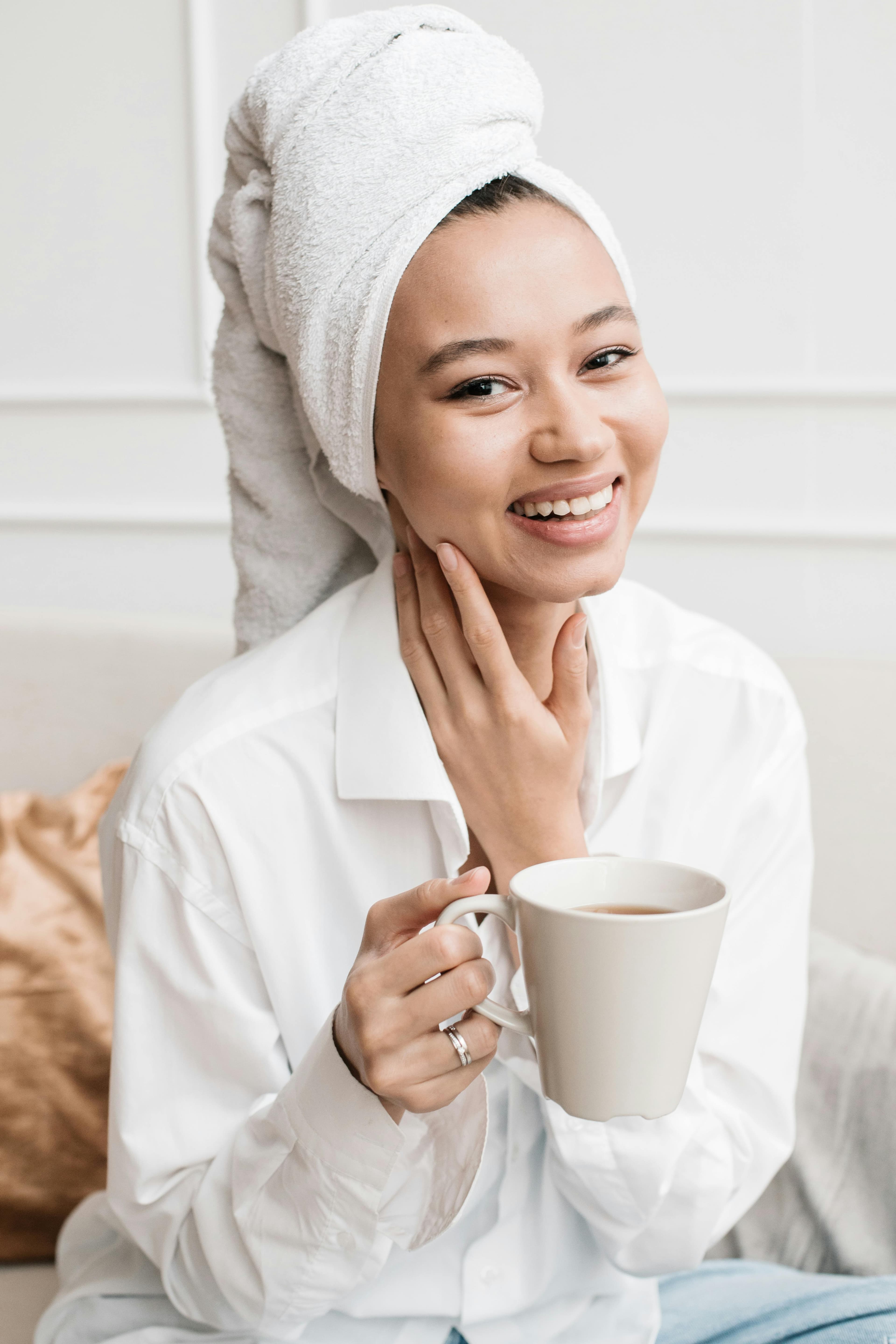 Happy woman with a towel on her head holding a cup, enjoying a cozy moment indoors.