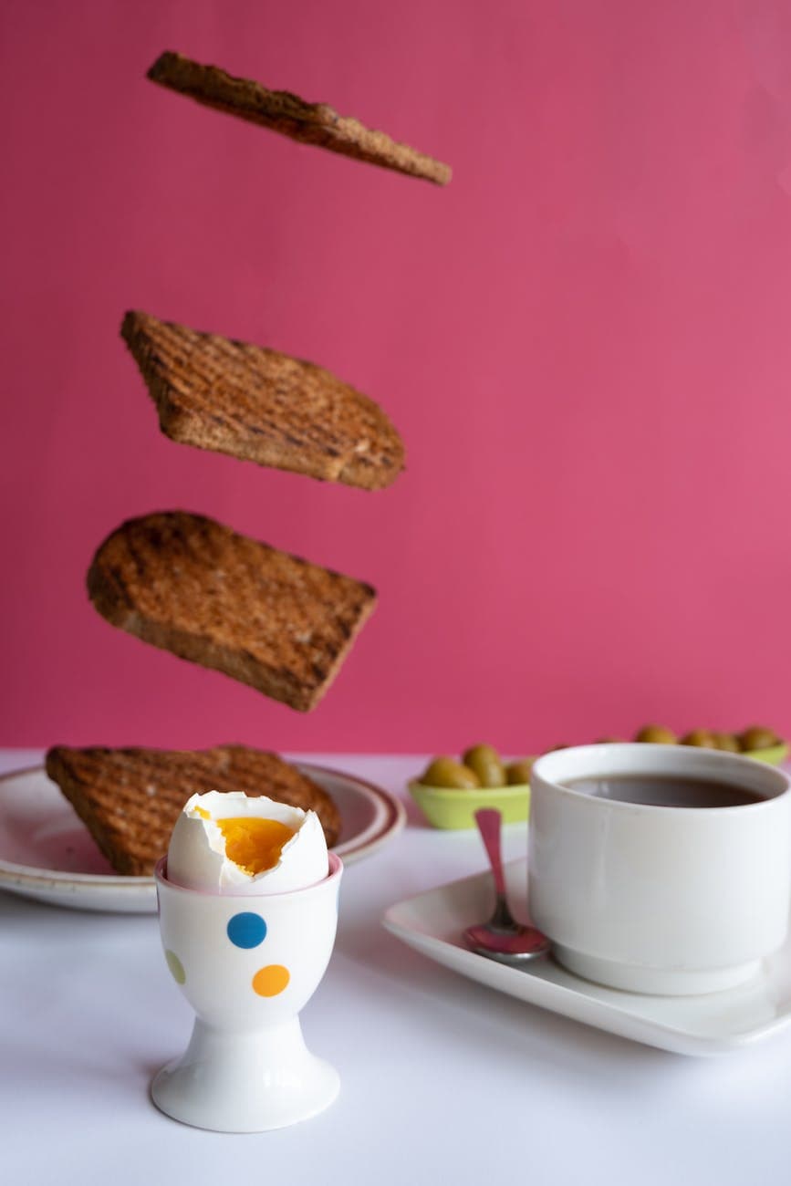 A playful breakfast setup with levitating toast, a soft-boiled egg, and coffee against a pink background.