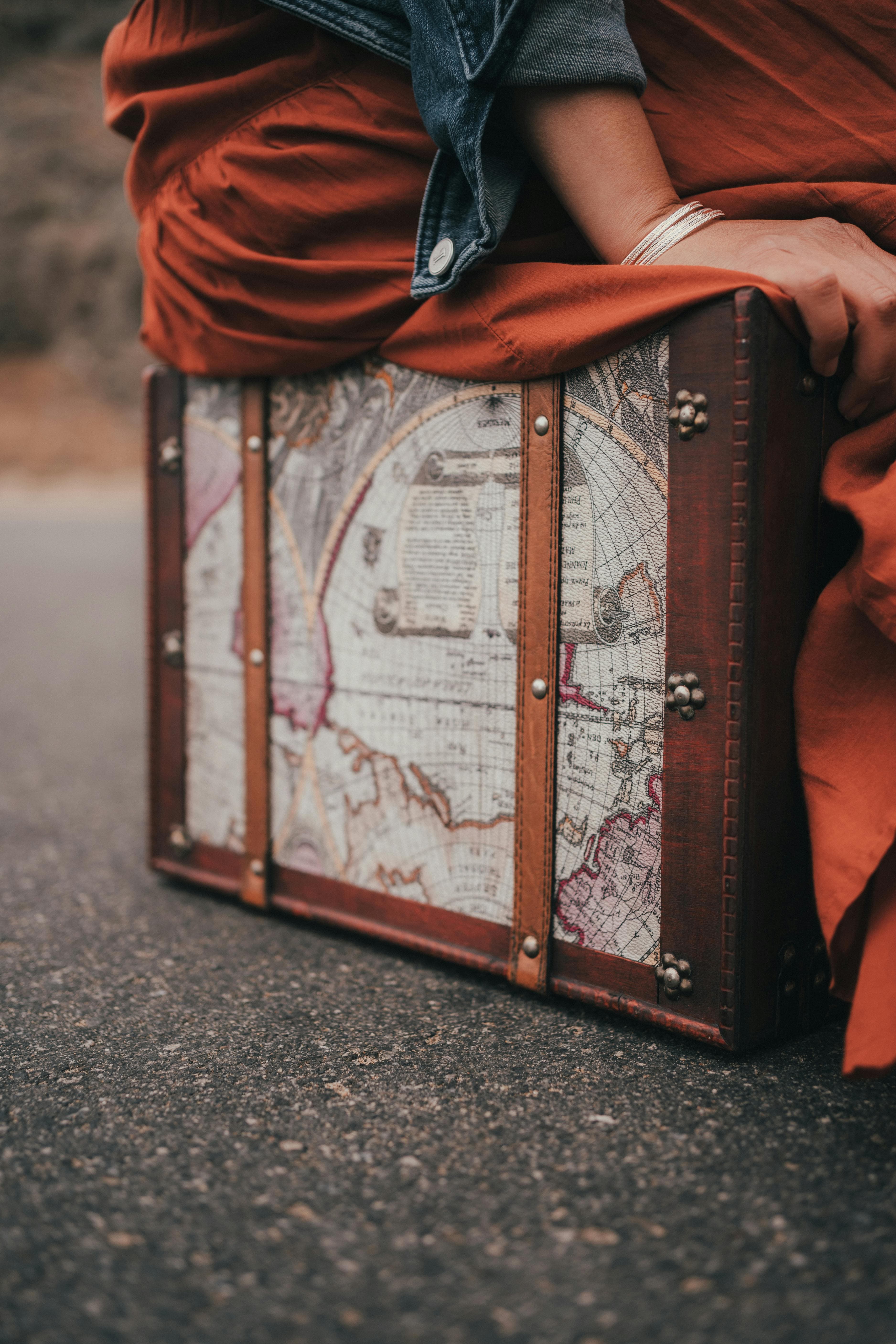 A woman sits on a vintage suitcase adorned with an old map design, outdoors.